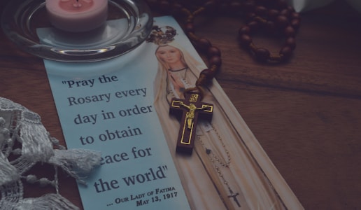A religious pamphlet features an illustration of Our Lady of Fatima with a quote about praying the rosary for world peace. A wooden rosary with a crucifix is placed on top of the pamphlet. A pink tea light candle sits on a clear glass holder nearby, and a piece of white lace fabric is partially visible at the edge.