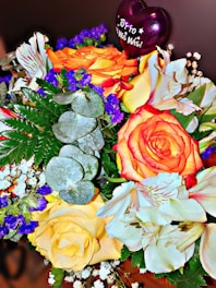 A vibrant bouquet featuring orange, yellow, and white flowers, accented by deep blue blossoms and green foliage. A purple heart-shaped balloon with 'Tú y yo un mes más!' adds a celebratory touch.