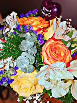 A vibrant bouquet featuring orange, yellow, and white flowers, accented by deep blue blossoms and green foliage. A purple heart-shaped balloon with 'Tú y yo un mes más!' adds a celebratory touch.