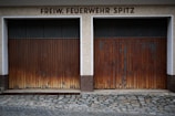 Two adjacent wooden garage doors with a weathered and worn appearance, set in a stone and concrete facade. Above the doors, the words 'FREIW. FEUERWEHR SPITZ' are displayed in bold, dark lettering. The ground is covered in cobblestones.