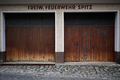Two adjacent wooden garage doors with a weathered and worn appearance, set in a stone and concrete facade. Above the doors, the words 'FREIW. FEUERWEHR SPITZ' are displayed in bold, dark lettering. The ground is covered in cobblestones.
