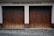 Two adjacent wooden garage doors with a weathered and worn appearance, set in a stone and concrete facade. Above the doors, the words 'FREIW. FEUERWEHR SPITZ' are displayed in bold, dark lettering. The ground is covered in cobblestones.
