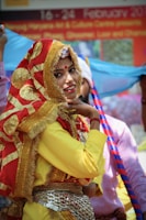 A person wearing traditional Indian attire with a red and gold embellished scarf covering their head. The outfit includes a vibrant yellow top and intricate jewelry, including a belt with coins. The background features a banner with text and other people in colorful clothing.