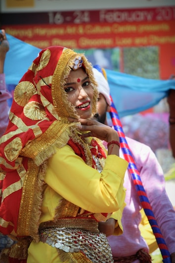 A person wearing traditional Indian attire with a red and gold embellished scarf covering their head. The outfit includes a vibrant yellow top and intricate jewelry, including a belt with coins. The background features a banner with text and other people in colorful clothing.