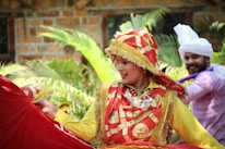 A person dressed in vibrant traditional attire with a red and gold theme, adorned with intricate patterns and designs. They are outdoors, surrounded by greenery and are engaged in what appears to be a lively dance or cultural performance. Another person in the background is dressed in white with a turban, contributing to the festive atmosphere.
