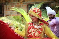 A person dressed in vibrant traditional attire with a red and gold theme, adorned with intricate patterns and designs. They are outdoors, surrounded by greenery and are engaged in what appears to be a lively dance or cultural performance. Another person in the background is dressed in white with a turban, contributing to the festive atmosphere.