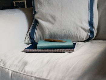 A neatly arranged stack on a white sofa, consisting of a spiral notebook on the bottom, a closed teal book in the middle, and a pen resting on top. The sofa has a pillow with blue stripes.