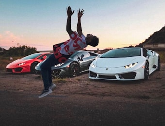 A person is performing a backflip in front of three luxury sports cars, with each car in a different color: red, black, and white. The setting is outdoors on a rough, unpaved surface, with a soft sunset or dawn sky providing a colorful backdrop.