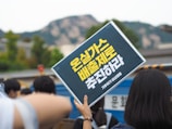 A group of people is gathered outdoors, holding up a sign with yellow text written in a non-Latin script. The background includes blurry trees and a mountain range, suggesting a public demonstration or rally.