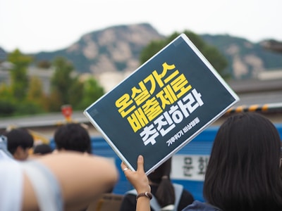 A group of people is gathered outdoors, holding up a sign with yellow text written in a non-Latin script. The background includes blurry trees and a mountain range, suggesting a public demonstration or rally.