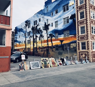 A mural covers the entire side of a multi-story building featuring a sunset scene with palm trees. In front of the building, various paintings and art pieces are displayed on the ground, likely part of a street art exhibition. The building has a sign that reads 'Venice Suites'. In the foreground, a car is partially visible.