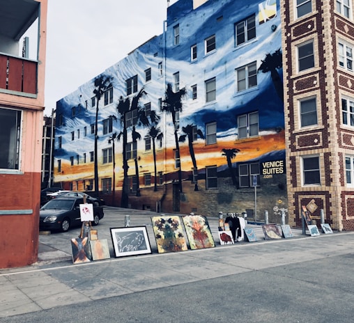 A mural covers the entire side of a multi-story building featuring a sunset scene with palm trees. In front of the building, various paintings and art pieces are displayed on the ground, likely part of a street art exhibition. The building has a sign that reads 'Venice Suites'. In the foreground, a car is partially visible.