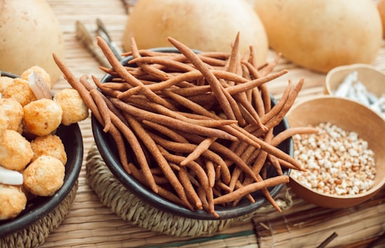 A collection of assorted snacks displayed in various bowls, including long, brown, finger-like snacks and round, golden, crispy balls. The arrangement is set on a textured, woven mat with additional small bowls containing seeds or grains. The background shows a soft focus with warm, earthy tones.