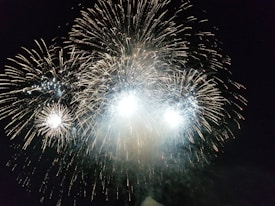Bright and colorful fireworks burst in the night sky, illuminating the darkness with dazzling patterns and sparkling lights. The display features large, radiant explosions accompanied by smaller, shimmering bursts around the edges.