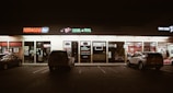 A strip mall storefront at night, featuring various businesses including a tobacco shop, nail and spa, and a donut shop. The area is dimly lit with neon signs, and a few cars are parked in front of the stores.