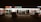 A strip mall storefront at night, featuring various businesses including a tobacco shop, nail and spa, and a donut shop. The area is dimly lit with neon signs, and a few cars are parked in front of the stores.
