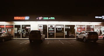 A strip mall storefront at night, featuring various businesses including a tobacco shop, nail and spa, and a donut shop. The area is dimly lit with neon signs, and a few cars are parked in front of the stores.