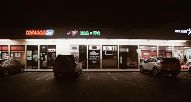 A strip mall storefront at night, featuring various businesses including a tobacco shop, nail and spa, and a donut shop. The area is dimly lit with neon signs, and a few cars are parked in front of the stores.