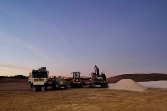 A construction site featuring various machinery including a truck, an excavator, and other construction vehicles. The ground is covered in dirt with a gravel pile in the foreground. The sky is a subtle gradient from light to darker blue, suggesting either early morning or late afternoon.