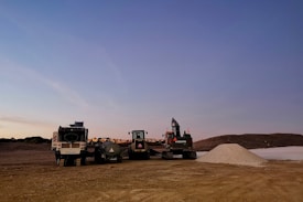 A construction site featuring various machinery including a truck, an excavator, and other construction vehicles. The ground is covered in dirt with a gravel pile in the foreground. The sky is a subtle gradient from light to darker blue, suggesting either early morning or late afternoon.