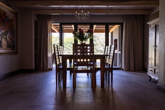 A dining room featuring a wooden table and six matching chairs with a flower arrangement placed centrally on the table. Large windows with drawn curtains line the back wall, allowing natural light to filter into the space. A chandelier hangs from the ceiling, and there is a painting on the left wall and an armoire or cabinet on the right.