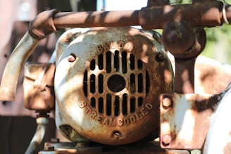 A close-up view of a rusty, weathered mechanical part with a fan cover. The surface features text indicating 'BALDOR' and 'STREAMCOOLED.' The metal exhibits significant rust and corrosion, giving it an aged appearance with patchy brown and orange colors. Sunlight casts shadows through the nearby foliage, enhancing the textured look of the rust.