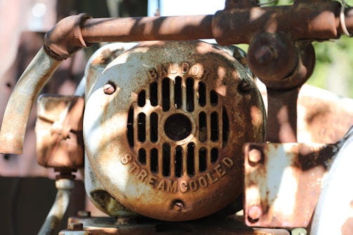 A close-up view of a rusty, weathered mechanical part with a fan cover. The surface features text indicating 'BALDOR' and 'STREAMCOOLED.' The metal exhibits significant rust and corrosion, giving it an aged appearance with patchy brown and orange colors. Sunlight casts shadows through the nearby foliage, enhancing the textured look of the rust.