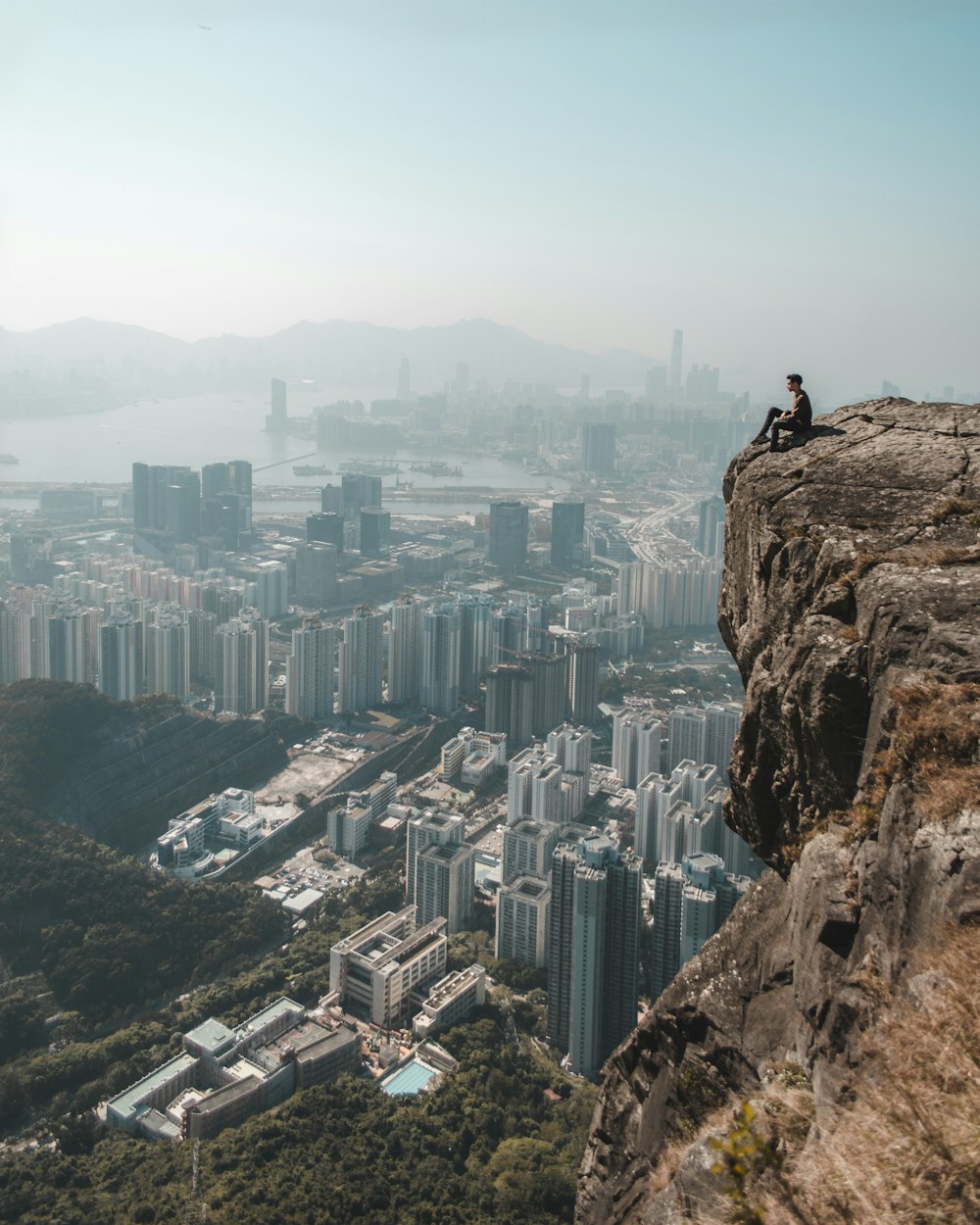 person sitting on cliff overlooking cityscape at daytime