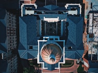 An aerial view of a large building with a prominent domed roof structure. The architecture features sharp, angular roofs with blue-gray shingles and a symmetrical layout. Surrounding structures include additional buildings and some construction work visible on one side.