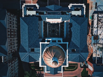 An aerial view of a large building with a prominent domed roof structure. The architecture features sharp, angular roofs with blue-gray shingles and a symmetrical layout. Surrounding structures include additional buildings and some construction work visible on one side.