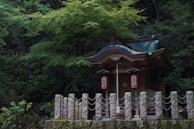 A small traditional Japanese shrine nestled in a lush forest setting. The shrine features a curved roof with intricate wooden carvings and hanging lanterns adorned with Japanese characters. Surrounded by dense green foliage and stone pillars connected by rope.