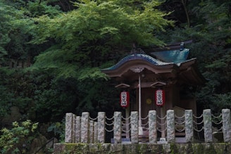 A small traditional Japanese shrine nestled in a lush forest setting. The shrine features a curved roof with intricate wooden carvings and hanging lanterns adorned with Japanese characters. Surrounded by dense green foliage and stone pillars connected by rope.