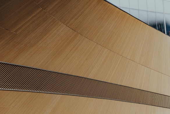 A modern architectural structure featuring sweeping curves and lines, with a textured wooden surface. A contrast is visible between the wooden elements and a portion of glass window at the top, reflecting light.
