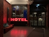 A dimly lit hotel lobby features a red neon sign spelling 'HOTEL' near a window. The wallpaper has a dark, intricate pattern, and there is a brown leather couch with turquoise throw pillows. Through the windows, bicycles and urban streetlights are visible outside. The lighting creates a warm and inviting atmosphere.