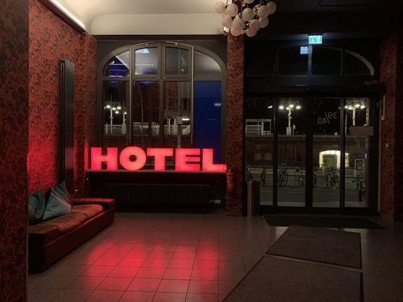 A dimly lit hotel lobby features a red neon sign spelling 'HOTEL' near a window. The wallpaper has a dark, intricate pattern, and there is a brown leather couch with turquoise throw pillows. Through the windows, bicycles and urban streetlights are visible outside. The lighting creates a warm and inviting atmosphere.