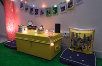 A cozy corner with a yellow trunk serving as a table, adorned with string lights and decorative items like a clock, books, and a glass bowl. Colorful framed photos hang above, and a prominent cushion with artistic designs rests on a padded seat. The floor is covered with artificial green grass, enhancing the warm ambiance.