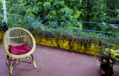 A wicker chair with a red cushion is positioned on a red-tiled balcony. The balcony is surrounded by lush green foliage and potted plants. A yellow wall with a blue railing runs the length of the balcony, providing a vibrant contrast to the greenery.