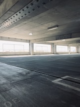 An empty parking garage with concrete floors and ceilings, featuring large windows that allow natural light to filter in. The structure is characterized by its industrial aesthetic, with visible steel beams and reinforced concrete.