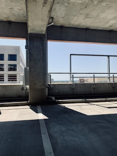 Inside a multi-story parking garage with concrete structures and a view of a distant building through an opening. Sunlight casts shadows on the concrete floor, and a metal railing can be seen in the background.