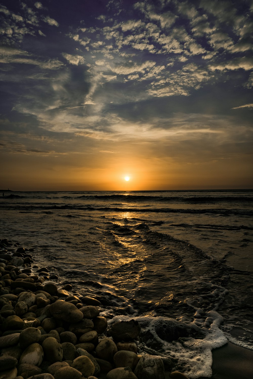 the sun is setting over the ocean with rocks in the foreground