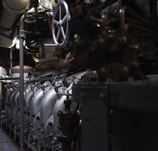 A complex array of industrial machinery and pipes with a large valve wheel dominates the scene. The machinery appears metallic and rugged, with various interconnected components.