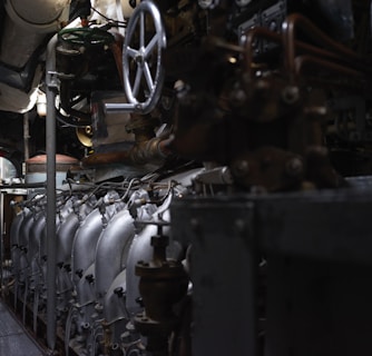 A complex array of industrial machinery and pipes with a large valve wheel dominates the scene. The machinery appears metallic and rugged, with various interconnected components.