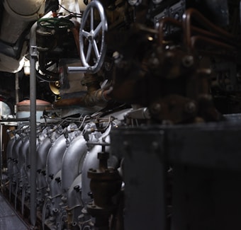 A complex array of industrial machinery and pipes with a large valve wheel dominates the scene. The machinery appears metallic and rugged, with various interconnected components.