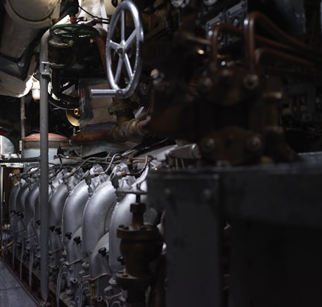 A complex array of industrial machinery and pipes with a large valve wheel dominates the scene. The machinery appears metallic and rugged, with various interconnected components.