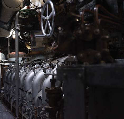 A complex array of industrial machinery and pipes with a large valve wheel dominates the scene. The machinery appears metallic and rugged, with various interconnected components.