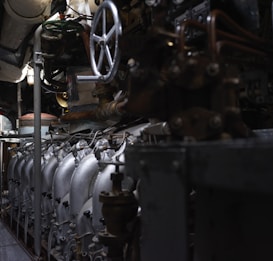 A complex array of industrial machinery and pipes with a large valve wheel dominates the scene. The machinery appears metallic and rugged, with various interconnected components.