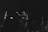 A group of people stand in a dimly lit space, some with their hands raised or outstretched, possibly engaging in an activity with enthusiasm or attentiveness. The setting has a muted, monochrome tone, enhancing the focus on the individuals' expressions and gestures.