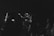 A group of people stand in a dimly lit space, some with their hands raised or outstretched, possibly engaging in an activity with enthusiasm or attentiveness. The setting has a muted, monochrome tone, enhancing the focus on the individuals' expressions and gestures.