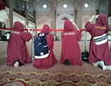 A group of people wearing maroon robes are kneeling on a carpeted floor inside an ornate building, possibly a mosque. They have their backs to the camera and are taking photos with their phones. The interior is decorated with intricate patterns and Arabic script on the walls.