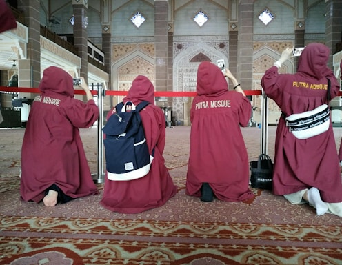 A group of people wearing maroon robes are kneeling on a carpeted floor inside an ornate building, possibly a mosque. They have their backs to the camera and are taking photos with their phones. The interior is decorated with intricate patterns and Arabic script on the walls.